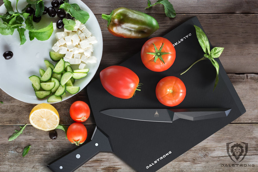 A black cutting board with full tomatoes on top of it next to a plate of green vegetables