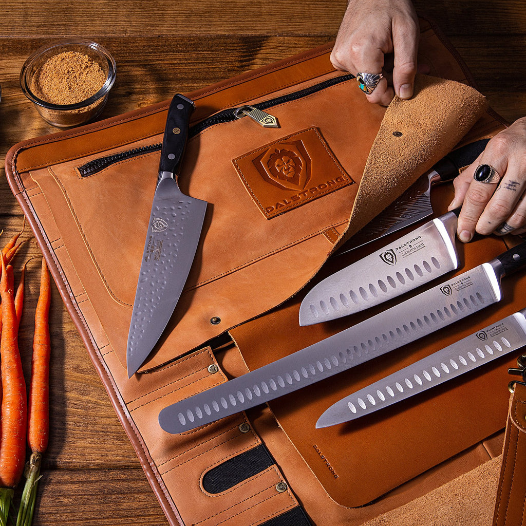 Four chef knives being placed into a brown leather knife roll with carrots next to it on a wooden table