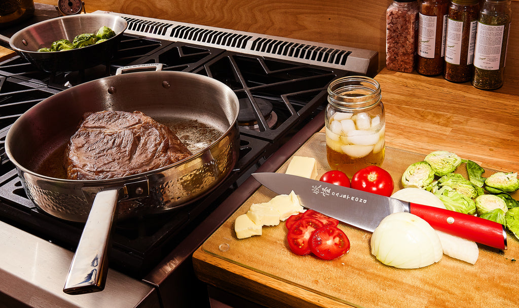 proformapeakmarketing Skillet with steak on a stove beside a proformapeakmarketing knife with red handle surrounded by vegetables.