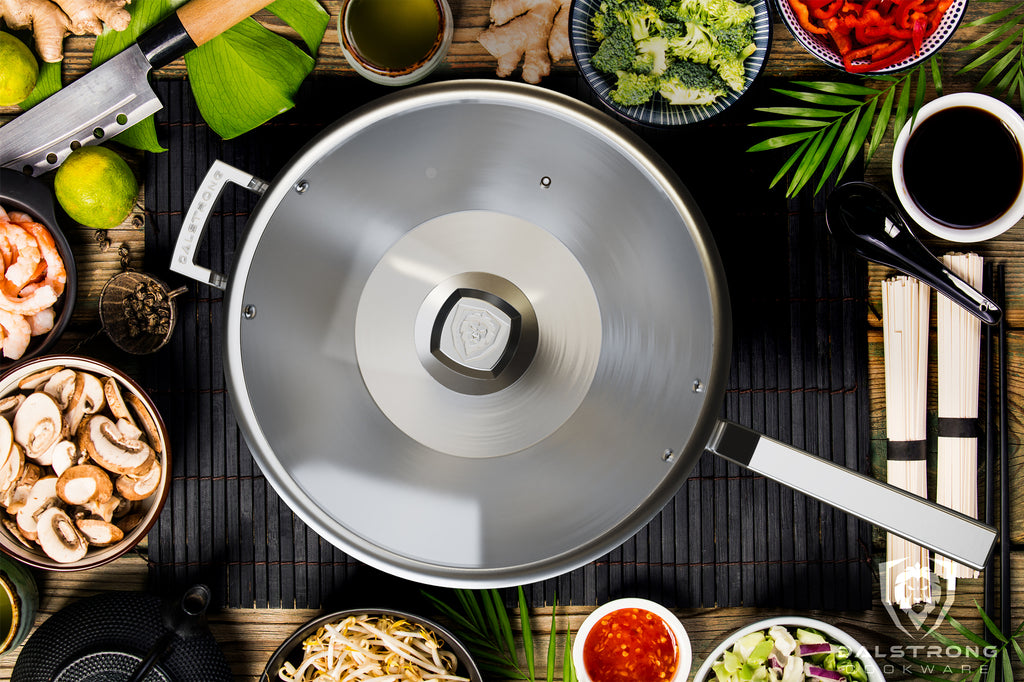 A wok with glass lid on a wooden surface surrounded by fresh vegetables