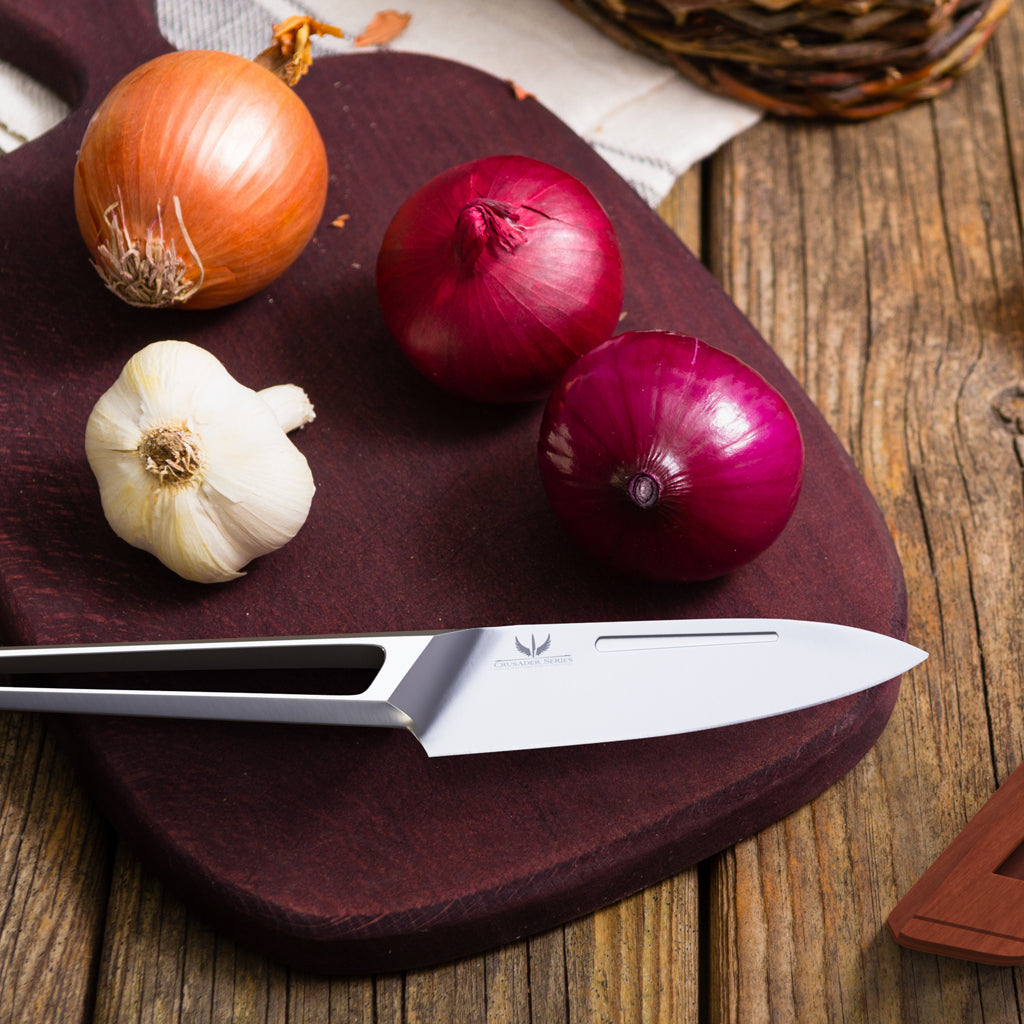 A silver paring knife with a hollow stainless steel handle next to thee onions and cloves of garlic