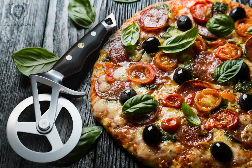 Pizza covered with toppings of olives tomatoes and basil on a grey table next to a pizza cutter