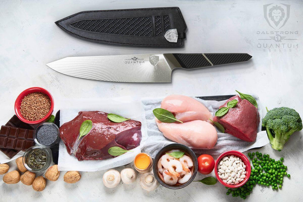 A variety of uncooked vegetables meat and poultry on a white surface next to a kiritsuke knife