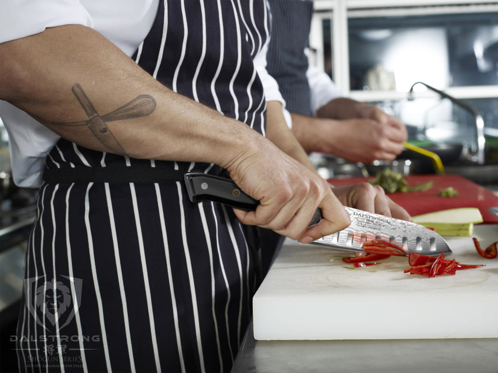 A chef with tattoos slices through a red pepper on a white cutting board with a Santoku