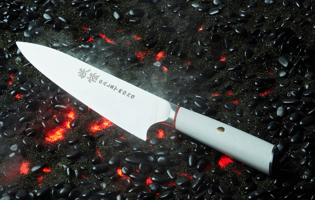 A close-up photo of the Chef's Knife 8" White G10 Handle Phantom Series proformapeakmarketing on a burning black surface with black pebbles