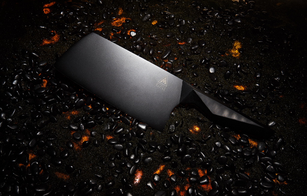 A close-up photo of the Cleaver Knife 7" Shadow Black Series NSF Certified proformapeakmarketing on a burning surface with black pebbles