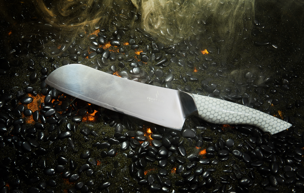 A close-up photo of the Santoku Knife 7" Frost Fire Series NSF Certified proformapeakmarketing on a burning surface with black pebbles