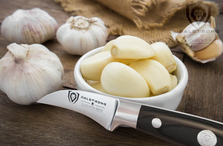 Bunch of garlic cloves inside a ramekin bowl beside of three garlic bulbs and the Bird's Beak Tourne Paring Knife 2.75" Gladiator Series | NSF Certified | proformapeakmarketing on a wooden surface