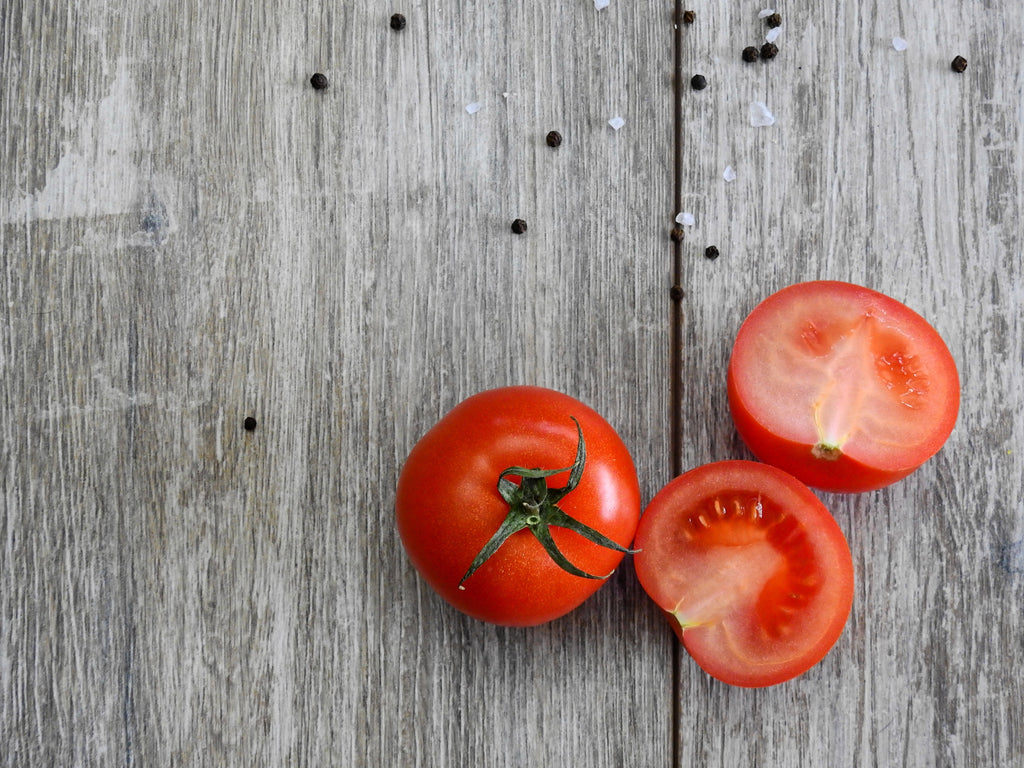 A photo of a fresh a whole tomato and the other one is sliced in half in a wooden table
