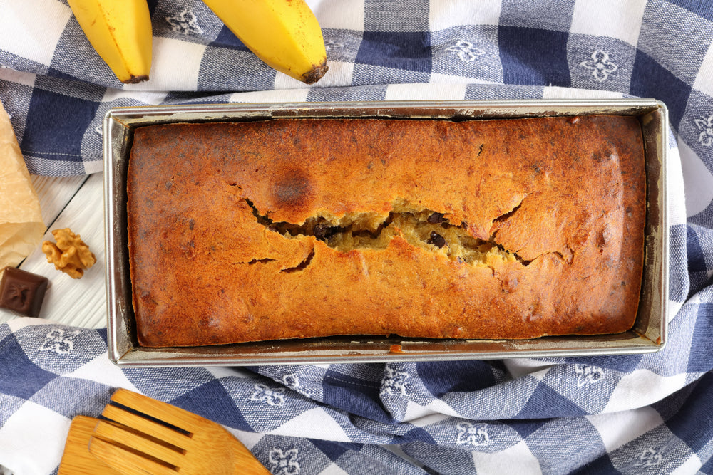 close-up of baked banana bread with walnuts and chocolate pieces in a loaf pan with kitchen towel, ripe bananas and ingredients on wooden table, authentic recipe, view from above