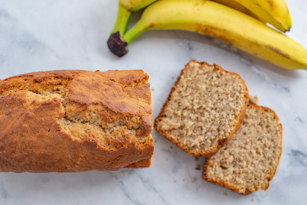 A banana bread loaf on a marble surface with two slices cut from the top beside two bananas