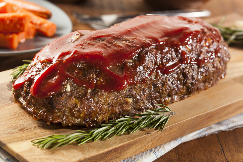 Meatloaf covered in sauce on a brown wooden cutting board with a plate of carrots in the background