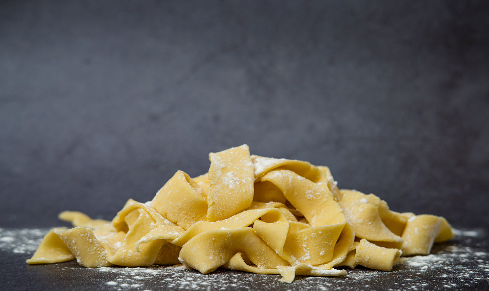 Fresh raw homemade uncooked gluten-free pappardelle pasta on dark table surface with flour on the table, very broad and flat pasta noodles