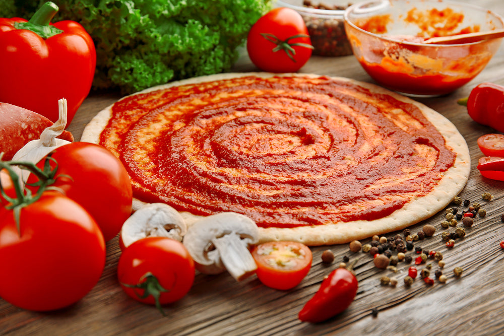 Uncooked Pizza base with tomato sauce spread across it and vegetables beside it on a wooden table