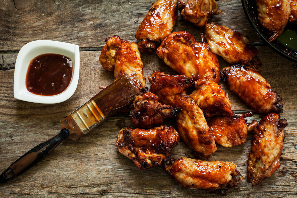 BBQ chicken wings on a wooden surface next to a jar of bbq sauce and a basting brush