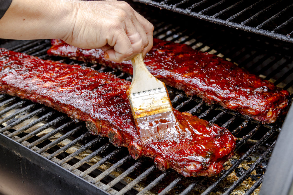 Two racks of pork bbq ribs sitting on grill with hand applying barbeque sauce with brush