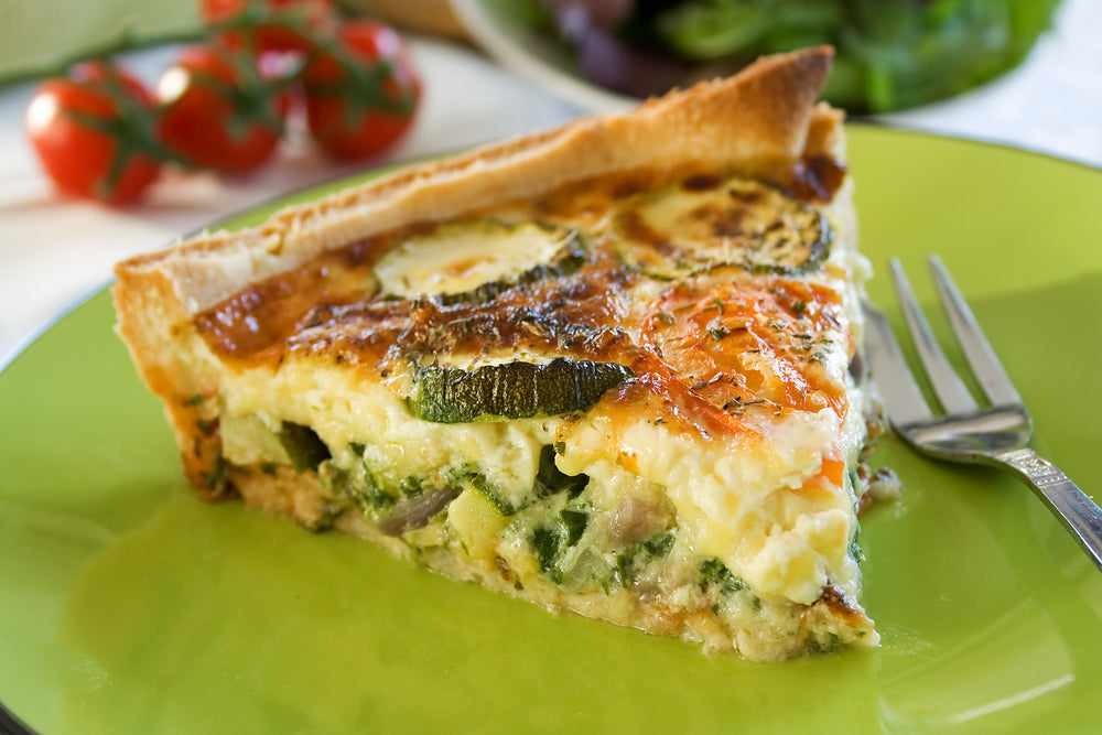A frittata slice on a green plate next to a fork with cherry tomatoes in the background