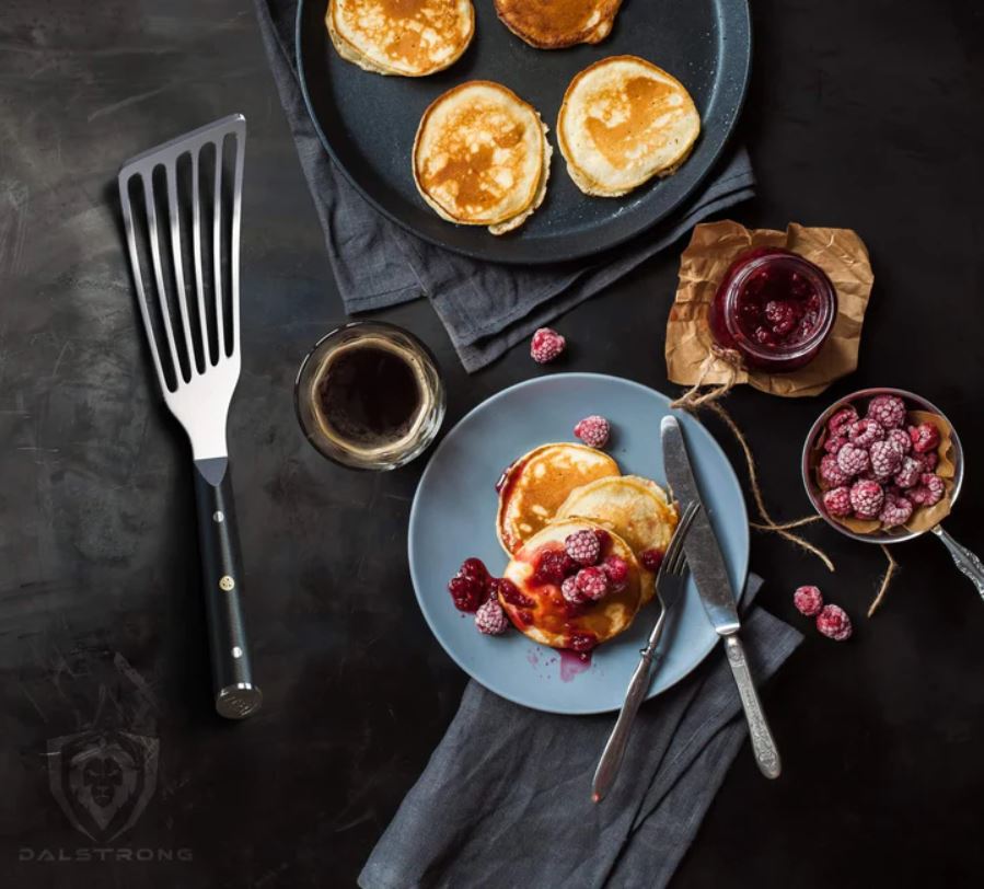 Professional Slotted Spatula on a black table next to a pan of freshly cooked pancakes, and plated pancake with raspberries and honey