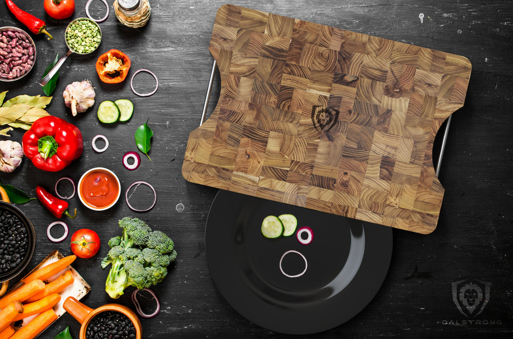 Teak Wood Cutting board surrounded by vegetables and a black dinner plate on a dark table